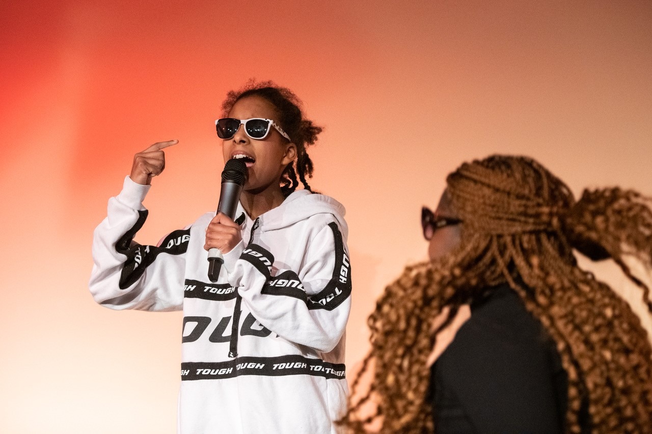 Zwei Mädchen, eine singend mit Mikro, eine im Profil und in Bewegung.