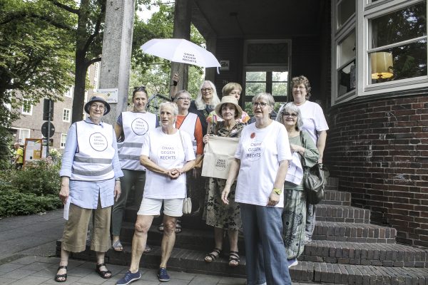 Zehn der OMAS GEGEN RECHTS aus Barmbek in T-Shirts oder Westen mit Logo. Eine Frau hält einen Regenschirm mit Logo der OMAS. Sie stehen auf einer Treppe vor dem KulturKlinker.